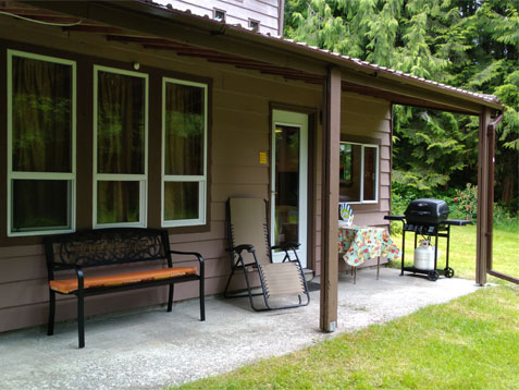 The Chown Riverside Cabin — covered porch with a bench, lounge chairs and a barbecue facing the lawn